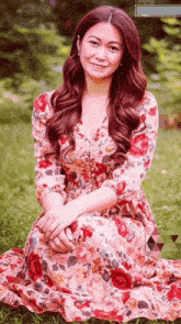 a woman in a floral dress sits in the grass with her hands folded