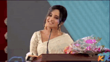 a woman stands at a podium with a bouquet of flowers in front of her and the letter k on the screen behind her