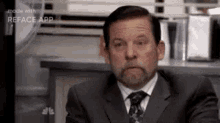 a man with a beard is wearing a suit and tie and sitting at a desk .