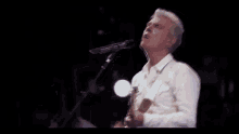 a man in a white shirt is singing into a microphone while playing a guitar .