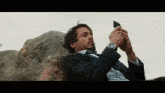 a man in a suit and tie is sitting on a rock taking a picture of himself with his cell phone .