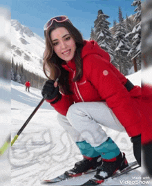 a woman in a red jacket is squatting down holding a ski pole