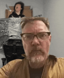 a man with glasses and a beard is taking a selfie with a picture of a man behind him