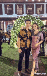 a man and a woman pose for a photo in front of a building with flowers in the background