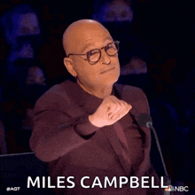 a bald man wearing glasses and a suit is giving a thumbs up in front of a microphone and says miles campbell