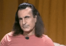 a man with long hair and a pink shirt is sitting in front of a brown curtain .