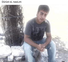 a man in a gray shirt is sitting on a rock next to a tree .