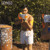 a shirtless man is holding a water gun in front of a barrel with skymed written on it