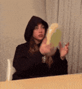 a woman in a black hoodie is sitting at a table with a sandwich in her hand