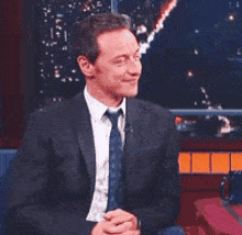 a man in a suit and tie is sitting in a chair with his hands folded and smiling .