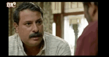 a man with a mustache is talking to another man on a tv screen .