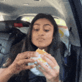 a woman sitting in a car eating a hamburger with a bag of wendy 's on the back seat