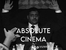 a man in a suit and tie is sitting at a table with his hands in the air and the words absolute cinema below him