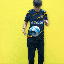a man wearing a aldi shirt is kicking a soccer ball on a yellow background