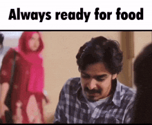 a man with a mustache is sitting at a table with a woman in a pink hijab standing behind him .