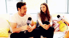 a man and woman are sitting on a couch talking into microphones .