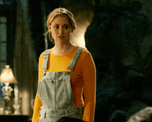 a woman wearing overalls and a yellow shirt is standing in a dark room