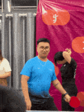 a man in a blue shirt is holding a ping pong paddle in front of a sign that says ' a '
