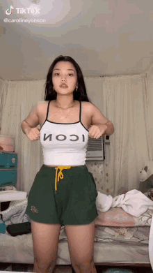 a woman in a white tank top and green shorts is standing next to a bed .