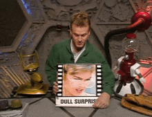 a man in a green jacket is holding a picture of a woman with glasses and a sign that says " dull surprise "