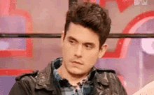 a man wearing a leather jacket and a plaid shirt is sitting in front of a red wall .
