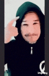 a person wearing a hooded jacket with a mustache is pointing at their head .