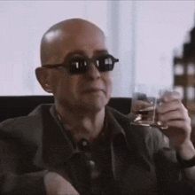 a bald man wearing sunglasses and holding a glass of whiskey