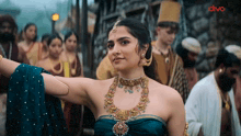 a woman in a blue dress and gold jewelry is standing in front of a crowd of people with the word dhvo on the bottom right