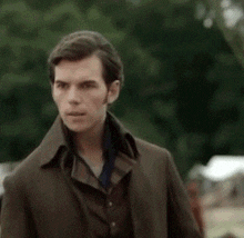 a man wearing a brown coat and a blue shirt is standing in front of trees