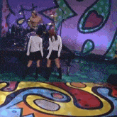 two women are standing on a stage with a man playing drums in the background