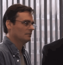 a man wearing glasses is standing in front of a window with blinds