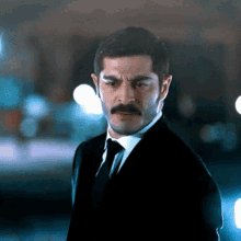 a man with a mustache wearing a suit and tie looks at the camera