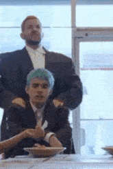 a man in a suit and tie is standing next to a man with blue hair at a table .