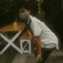 a man wearing a mask is squatting down in front of a wooden table and bench .