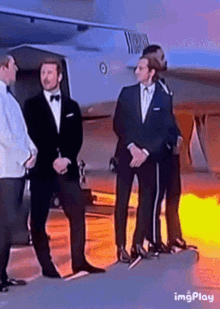 a group of men in tuxedos are standing in front of a plane with a flame coming out of it
