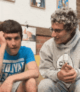 a man in a blue shirt that says 70yat sits next to another man in a gray hoodie