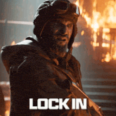 a man with a beard wearing a helmet and goggles stands in front of a burning building with the words lock in below him