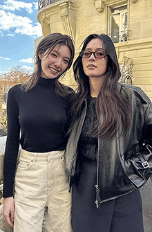 two women are posing for a picture in front of a building . one of the women is wearing glasses and a leather jacket .