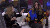two women are sitting on a couch holding wine glasses and talking .