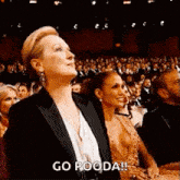 a woman in a tuxedo is sitting in front of a crowd and says `` go pooda '' .