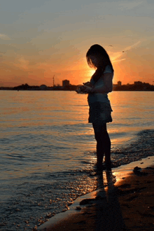 a woman standing on a beach looking at her phone at sunset