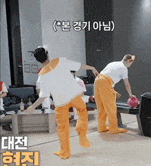 a man in orange pants is throwing a bowling ball while another man in white pants watches