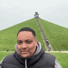 a man in a black vest stands in front of a large grassy hill