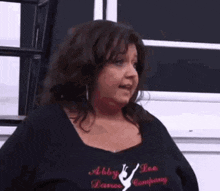 a woman wearing a black shirt that says abby lee dance company is standing in front of a window .