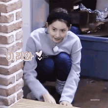 a woman is squatting down in front of a brick wall while wearing a headband and a blue sweater .
