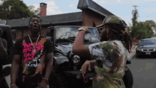 two men are standing in front of a jeep and one is wearing a t-shirt that says ' baddies ' on it