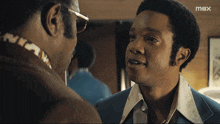 a man in a blue suit talks to another man with a max logo in the corner