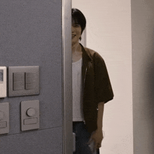 a man in a brown jacket stands in a doorway with a light switch on the wall behind him