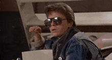 a man wearing sunglasses and a denim jacket is holding a piece of paper in his hand .