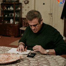 a man sits at a table using a calculator with a star logo on the wall behind him